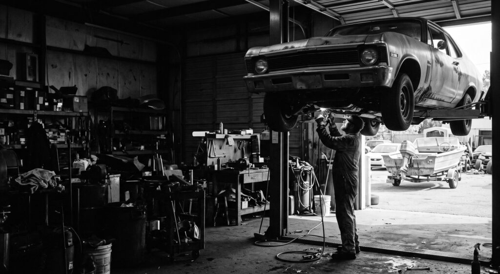 A mechanic working under a car on a lift with a boat on a trailer outside the bay